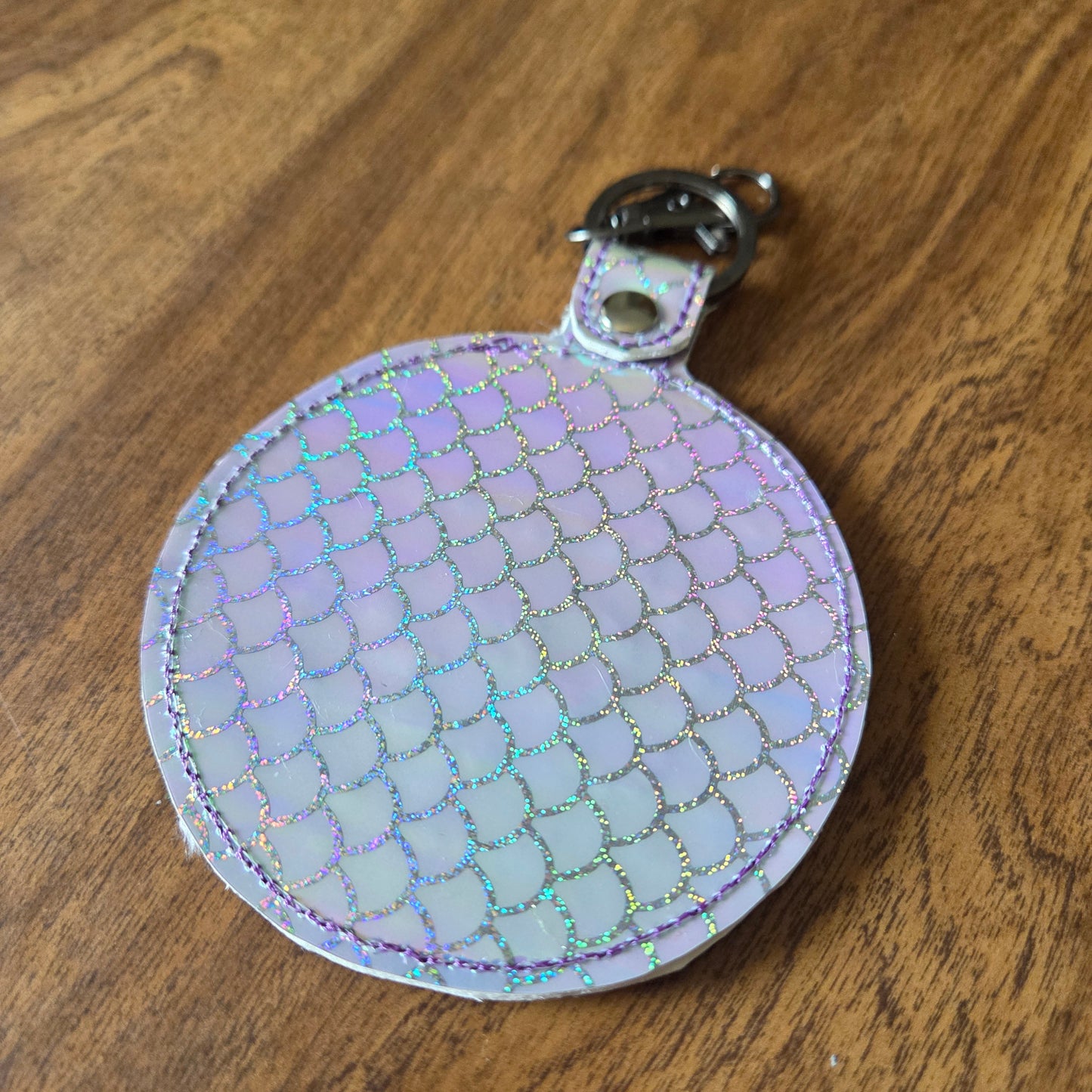 A small round iridescent keychain pouch with a scale-like pattern, attached to a silver metal clasp, resting on a brown wooden surface.