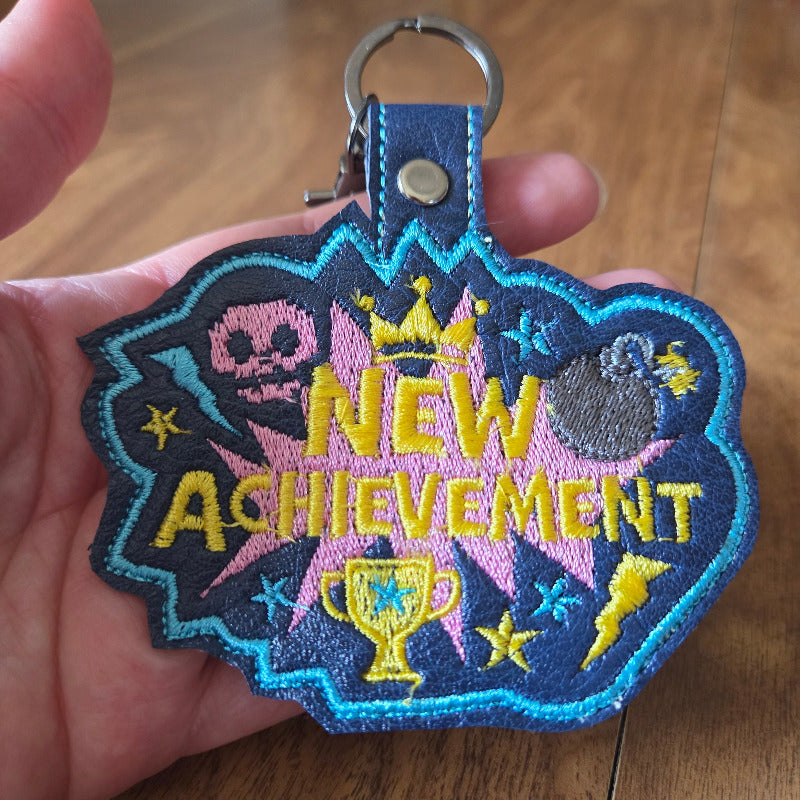 A hand holding a decorative embroidered keychain patch that reads 'New Achievement' in yellow text over pink and blue starburst shapes, with a gray trophy, yellow lightning bolt, skull, and stars. The patch is attached to a blue strap with a silver metal key ring, shown against a wooden surface.