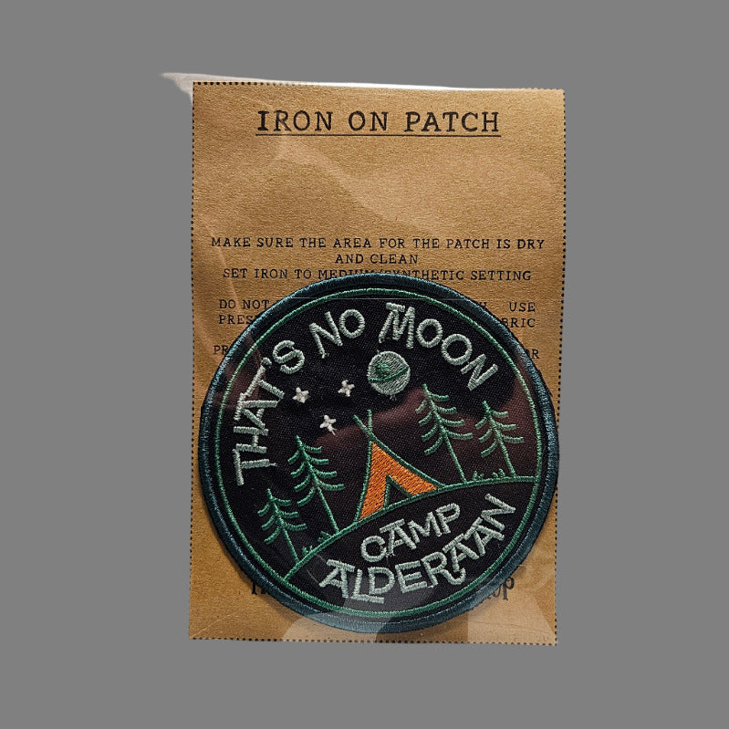 Round embroidered patch with text and graphics on a cardboard backing, placed on a textured surface.