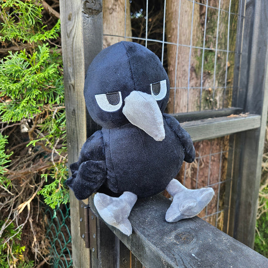 Plush toy resembling a black and white creature on a wooden surface with greenery in the background