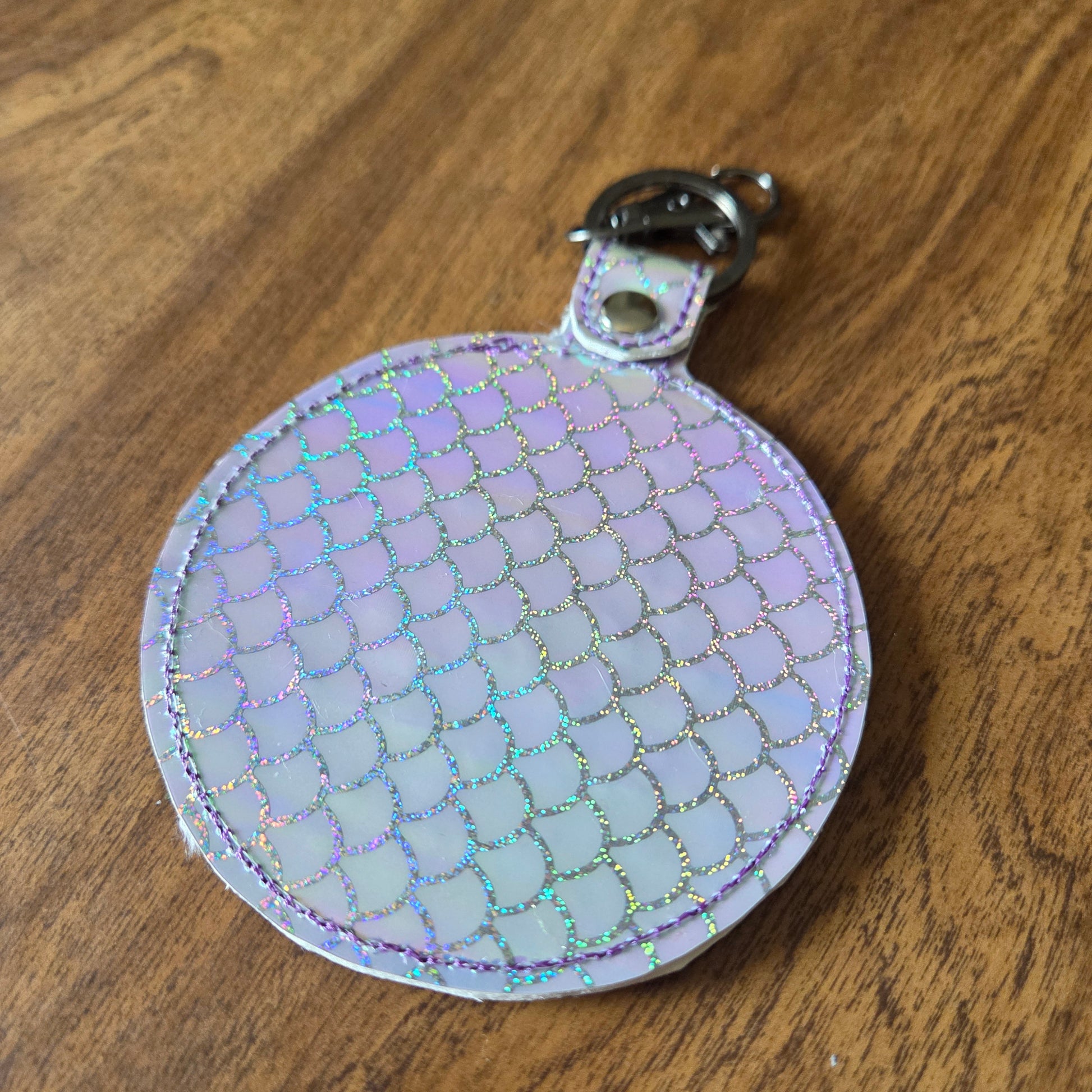 A small round iridescent keychain pouch with a scale-like pattern, attached to a silver metal clasp, resting on a brown wooden surface.