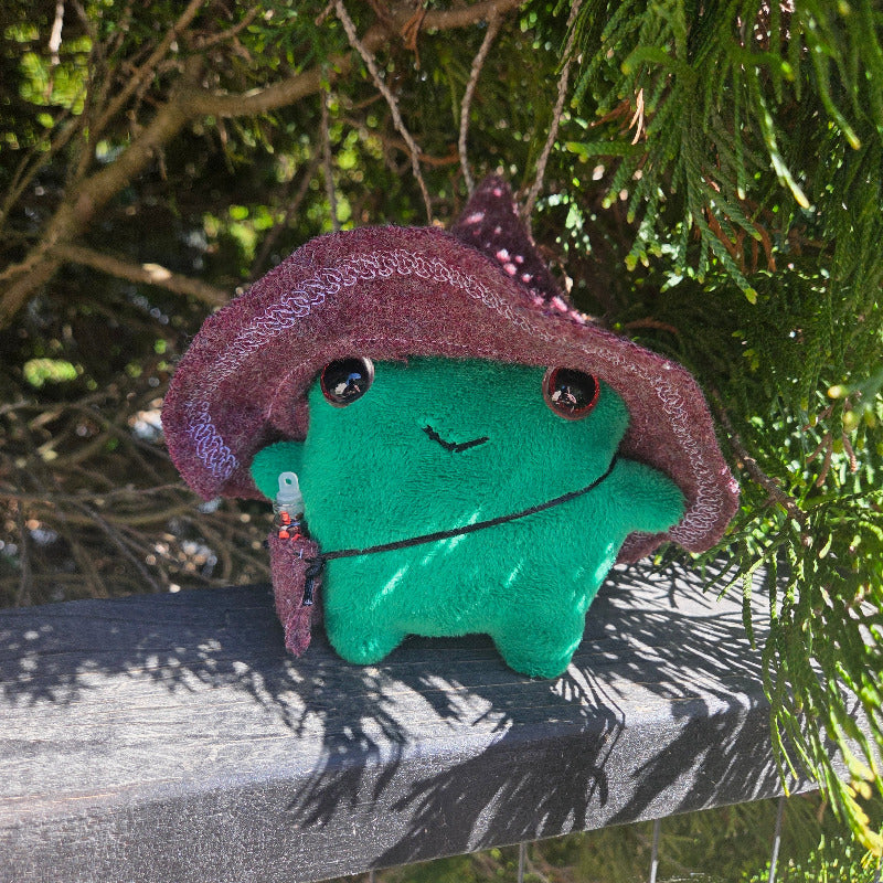 Plush toy resembling a green creature with a brown hat against a natural background