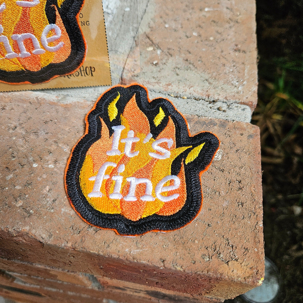 Flame-themed patch with 'it's fine' text on a brick surface