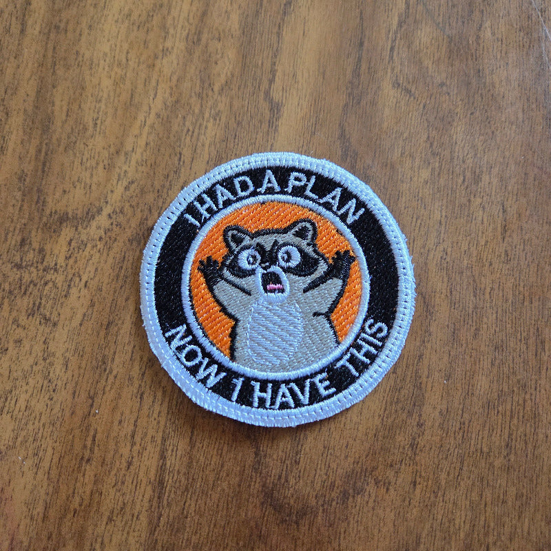 A circular embroidered patch featuring a raccoon with the text 'HAD A PLAN I NOW LIVE THIS' on a wooden surface.