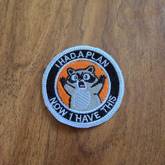 A circular embroidered patch featuring a raccoon with the text 'HAD A PLAN I NOW LIVE THIS' on a wooden surface.