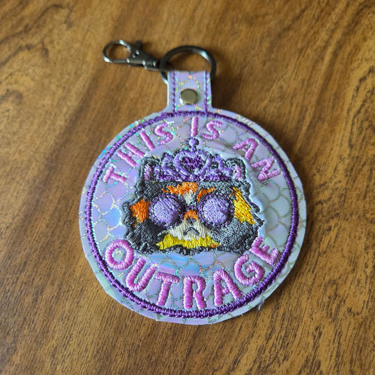 A round embroidered patch with a metal clasp, featuring the text 'THIS IS AN OUTRAGE' in purple around a central embroidered design of a cat wearing sunglasses, displayed on a wooden surface.