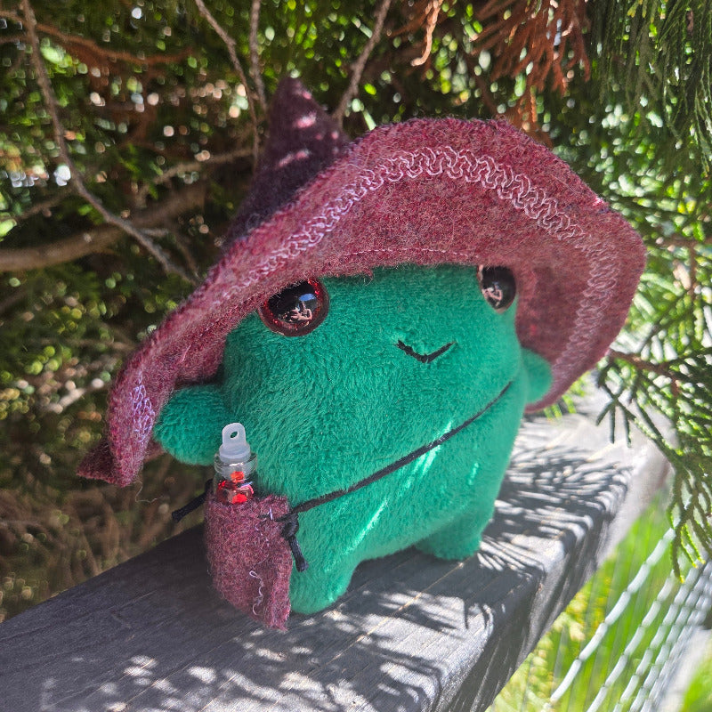 Plush toy resembling a green frog with a purple cape hanging from a tree branch.