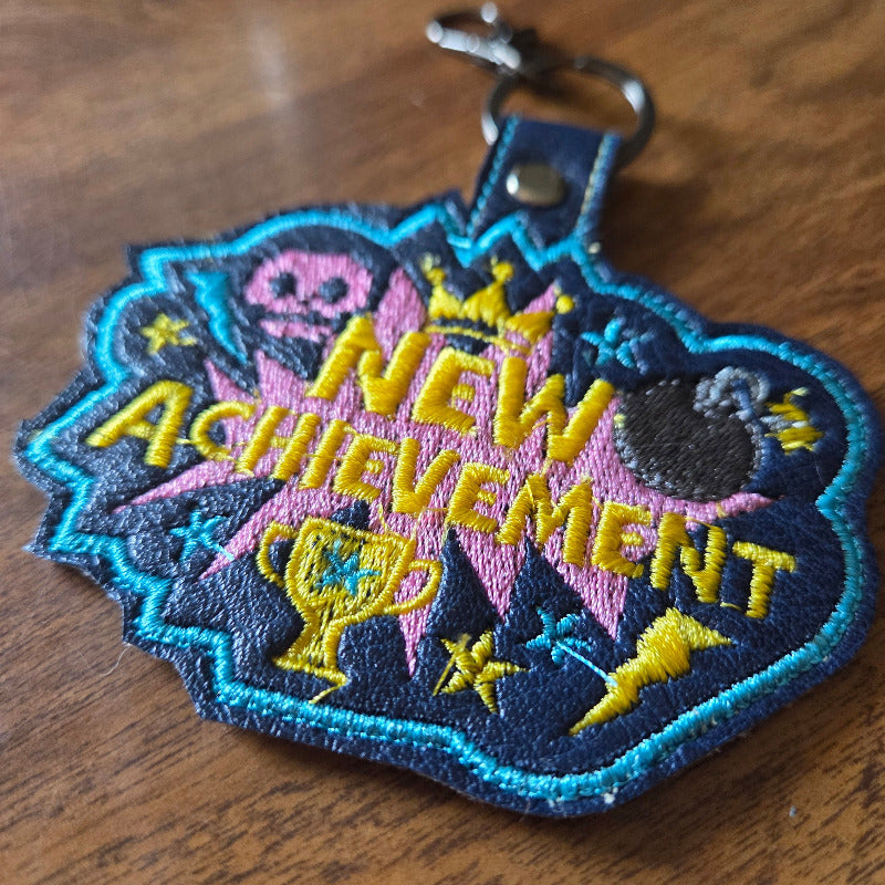 An embroidered patch keychain featuring the words 'NEW ACHIEVEMENT' in yellow, with pink and blue starburst shapes, stars, a trophy, and a skull on a dark background, attached to a metal key ring and photographed on a wooden surface.
