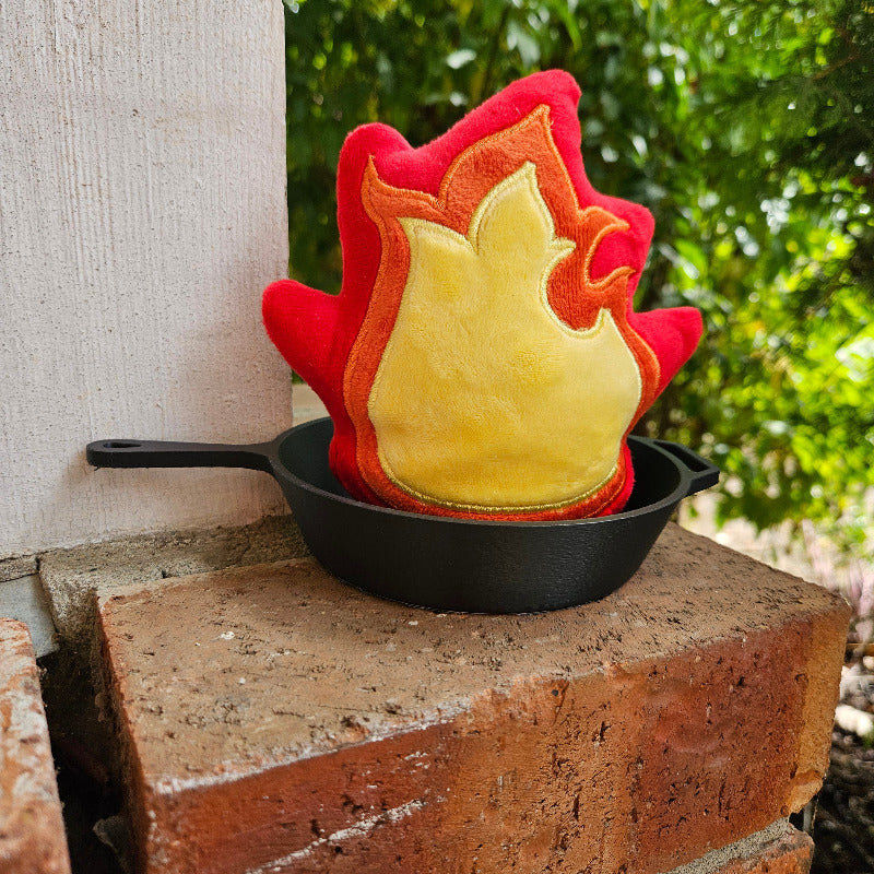 Flame-shaped potholder on a black cast iron skillet against a brick wall with greenery in the background