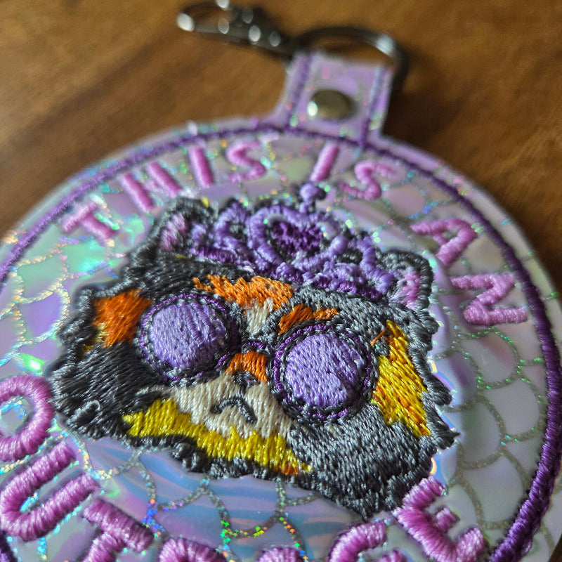 A close-up of a round embroidered patch or badge with a purple and lavender background, featuring a stylized face in black, purple, orange, and yellow embroidery. The patch has iridescent sequin detailing and is photographed on a wooden surface.