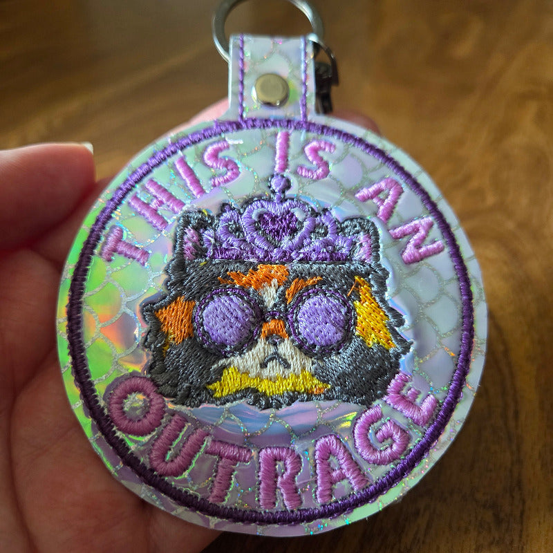 A close-up of a hand holding a round embroidered keychain or bag charm. The design features a purple and black skull with orange and yellow accents, surrounded by the text 'THIS IS AN OUTRAGE' in purple embroidery on a holographic background.