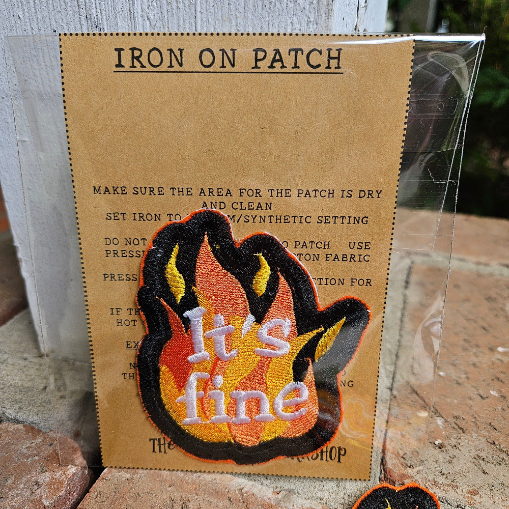 Iron-on patch with flame design and 'it's fine' text on a cardboard backing.