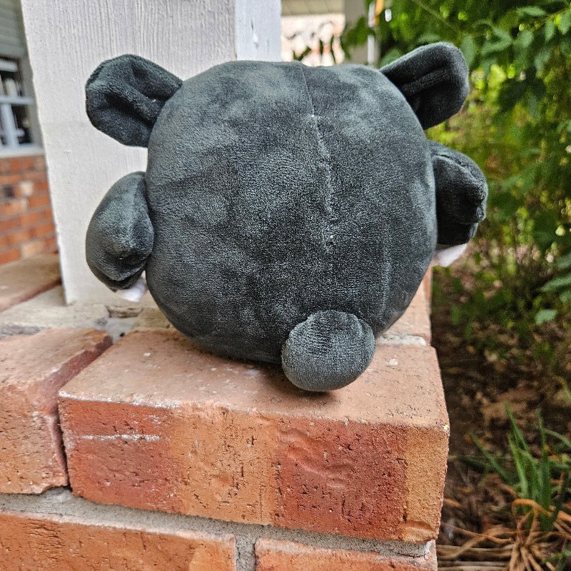 Gray plush toy resembling a raccoon on a brick surface with greenery in the background