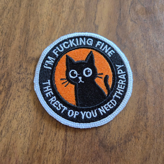 A circular embroidered patch featuring a black cat illustration on an orange background, surrounded by the text 'I'M FUCKING FINE THE REST OF MY LIFE YOU NEED THERAPY' in white lettering, placed on a wooden surface.