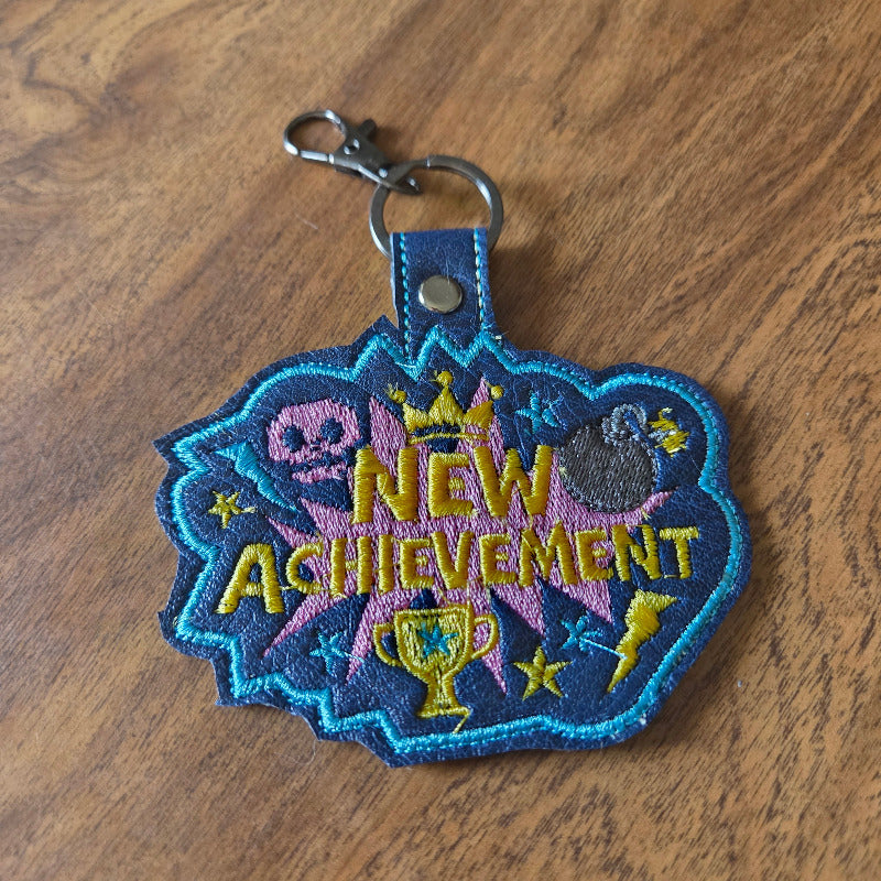 An embroidered patch keychain featuring a blue, purple, and yellow 'New Achievement' design with a silver clasp, resting on a wooden surface.